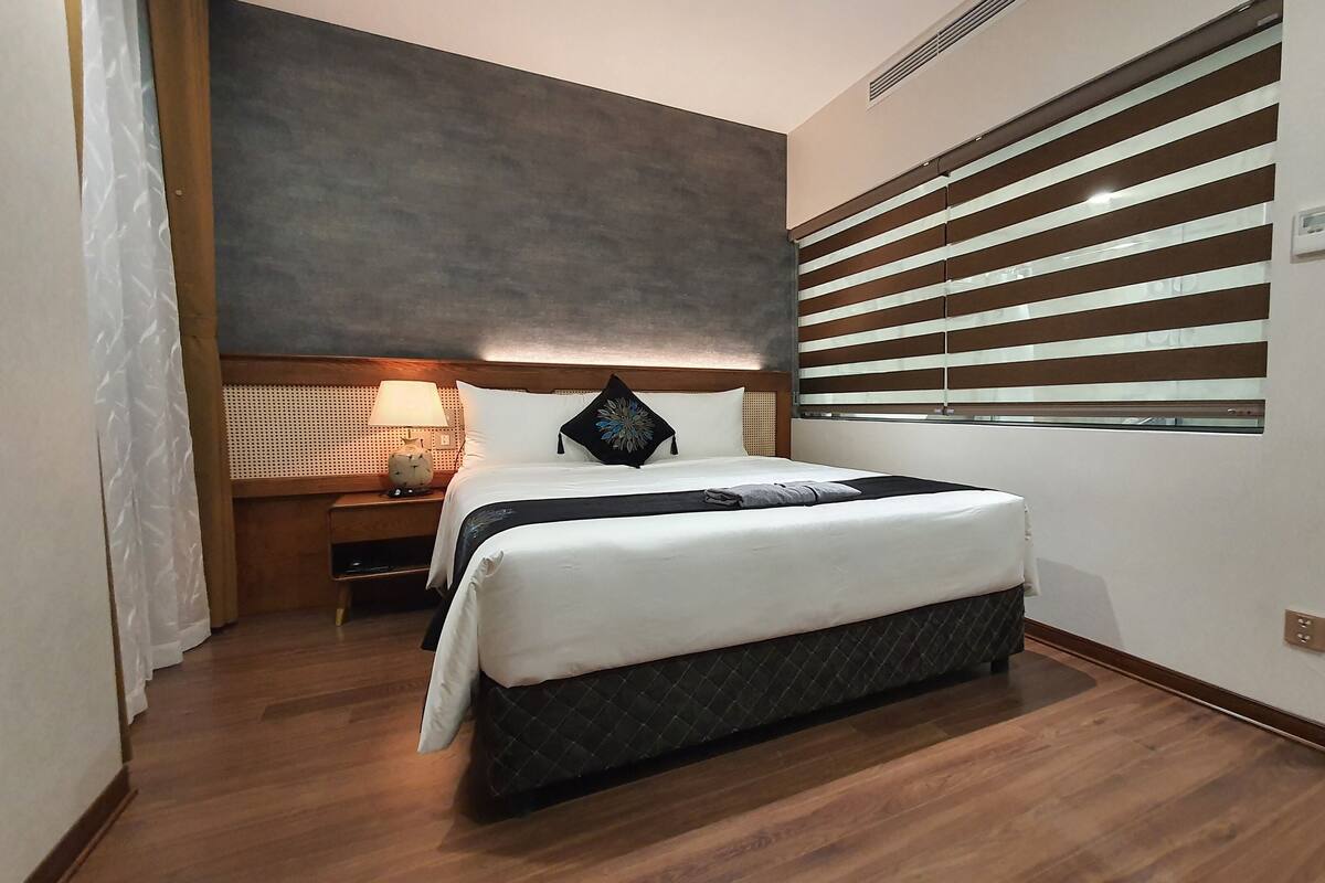 Una habitación de hotel con una cama grande, una mesita de noche con lámpara y una ventana con persianas.