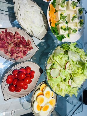 Ein Tisch mit verschiedenen Speisen, darunter Gurkenscheiben, Speck, Kirschtomaten, Feldsalat mit Radieschen, Tofu mit Gemüse und Eierscheiben.