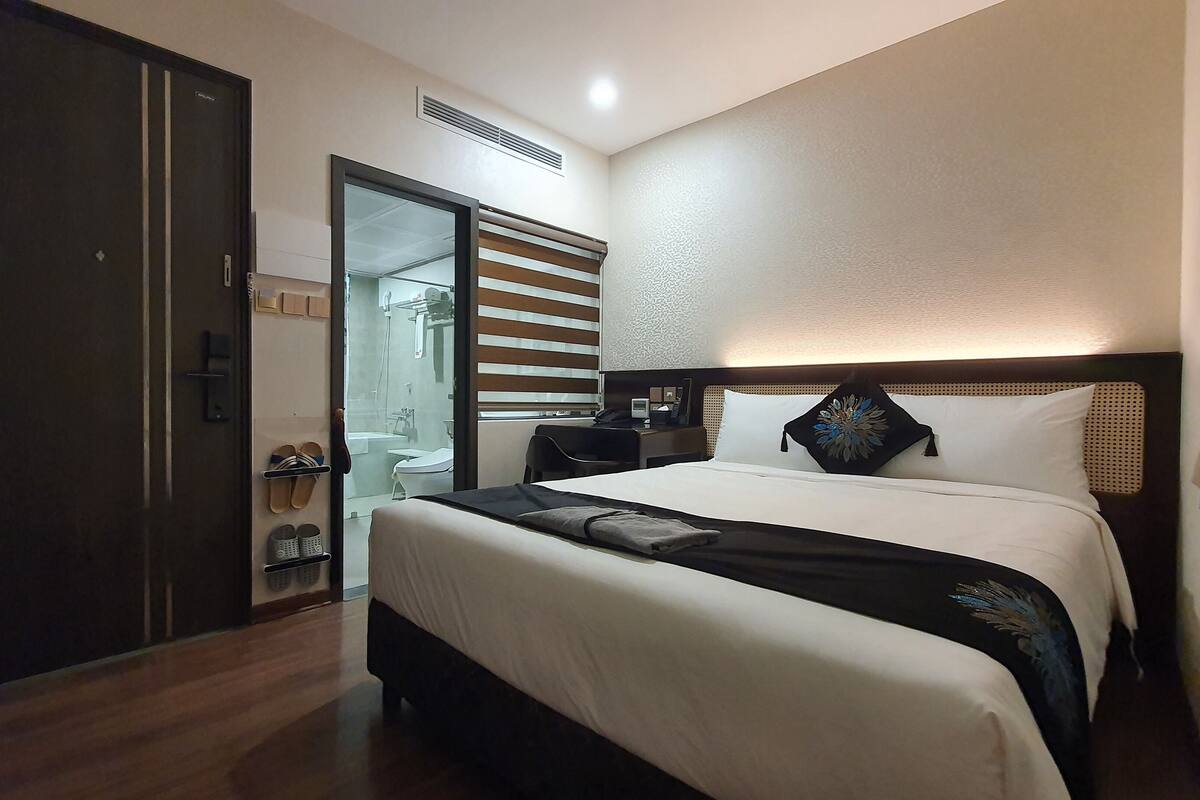 Habitación de hotel con cama, escritorio, armario y baño visible a través de una puerta de cristal.