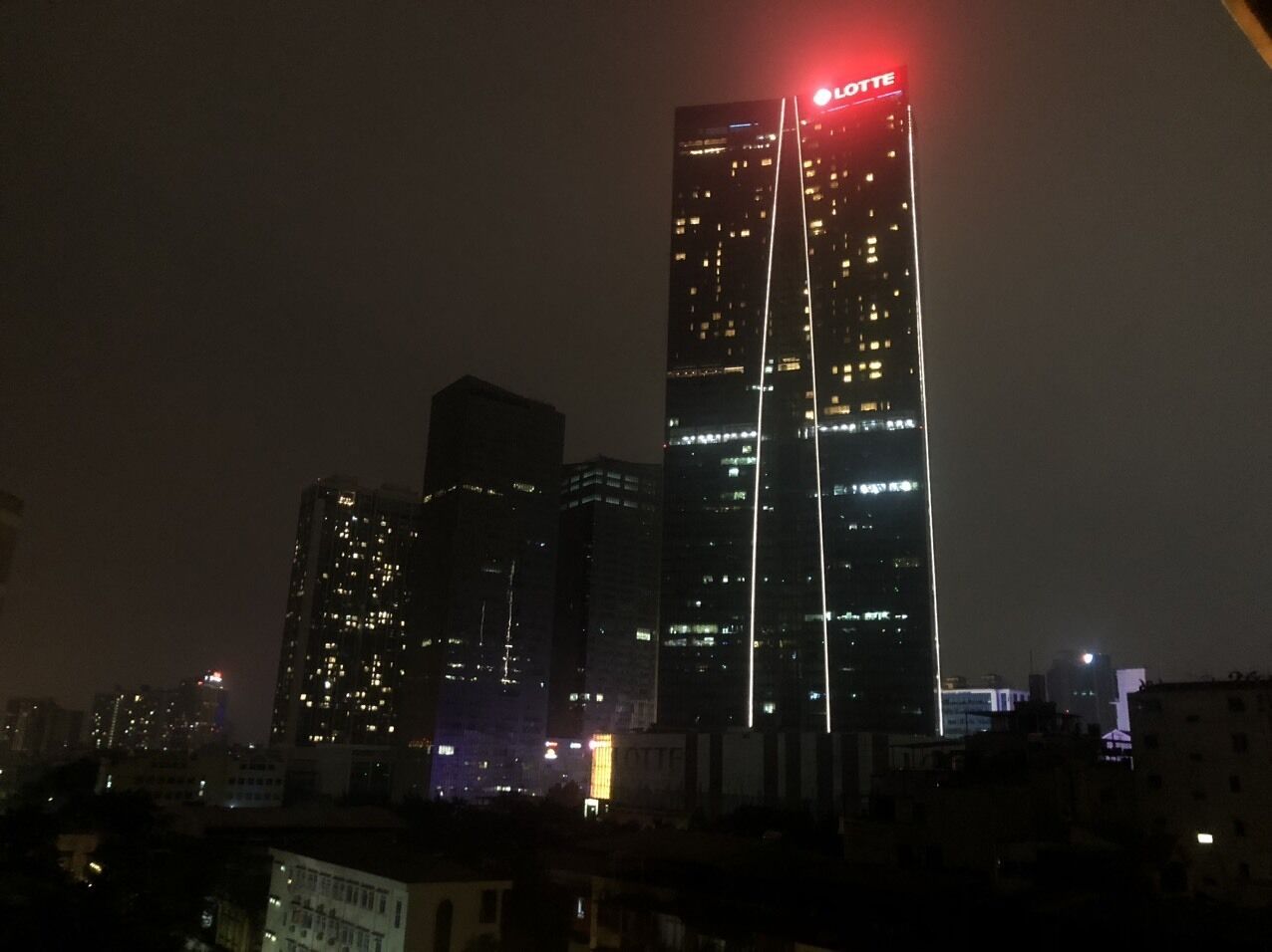 Un paisaje urbano nocturno con un rascacielos destacado, ventanas iluminadas y una luz roja en la parte superior.