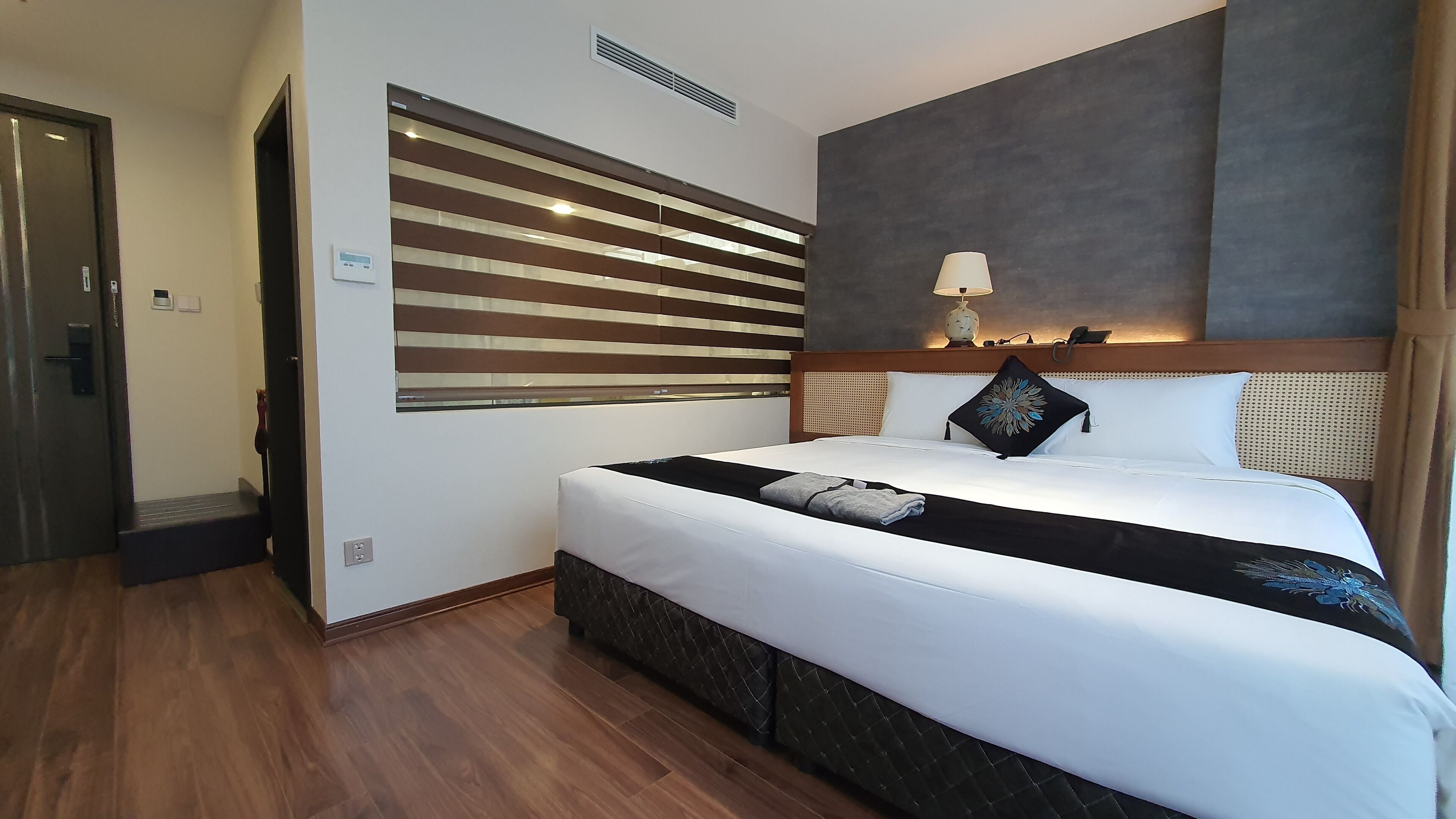 A hotel room with a large bed, a headboard with a light fixture, a wall-mounted air vent, and a door with a handle.
