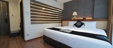 A hotel room with a large bed, a headboard with a light fixture, a wall-mounted air vent, and a door with a handle.