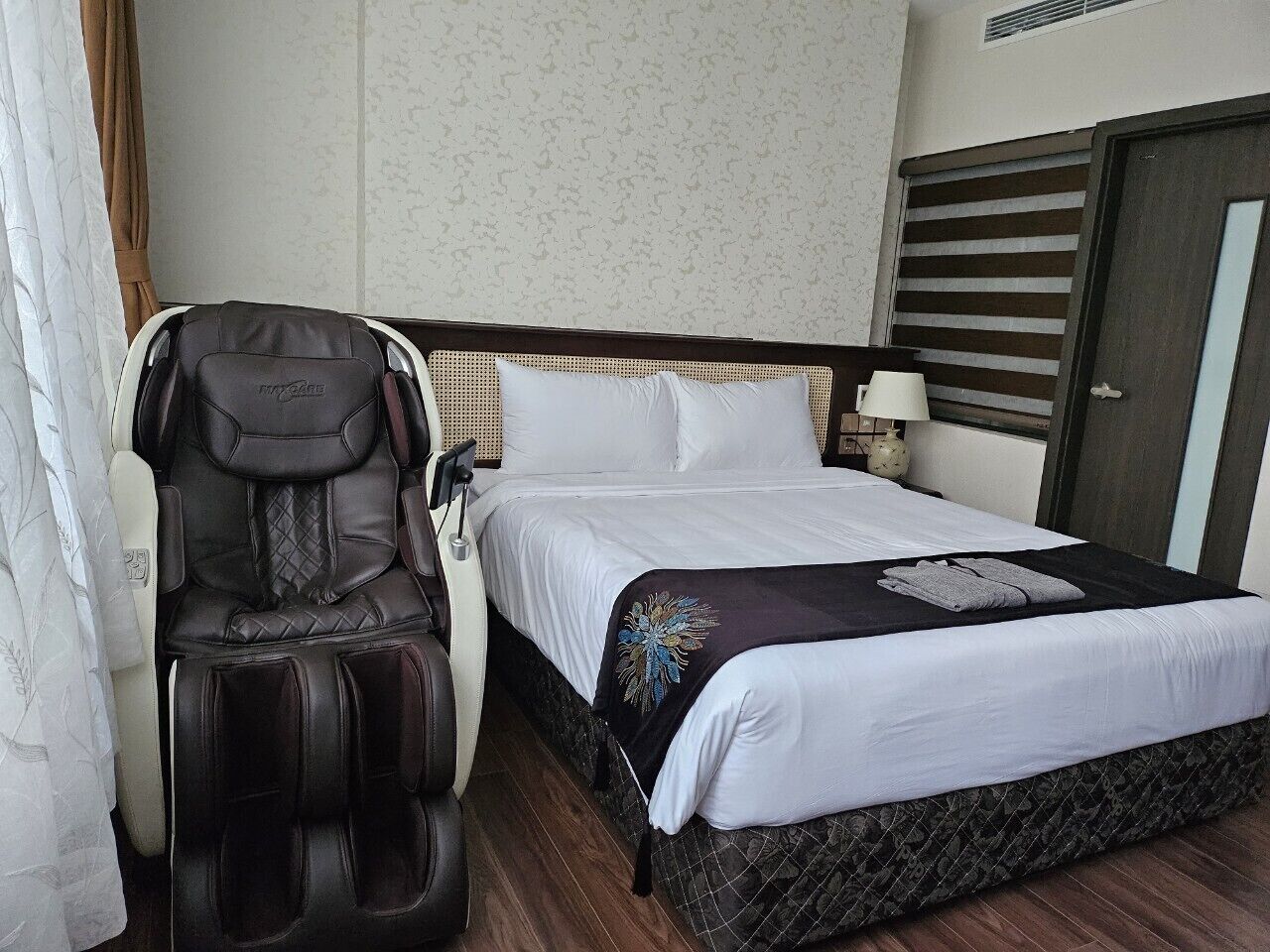 A hotel room with a bed, a massage chair, and a nightstand with a lamp.