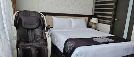A hotel room with a bed, a massage chair, and a nightstand with a lamp.