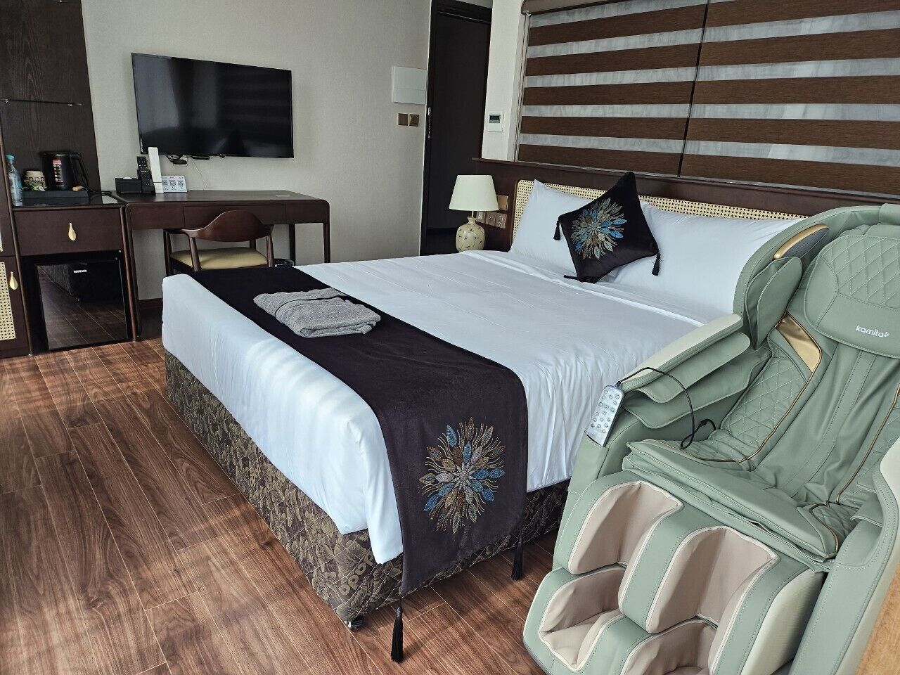 A hotel room with a bed, a massage chair, a desk with a TV, and a minibar.