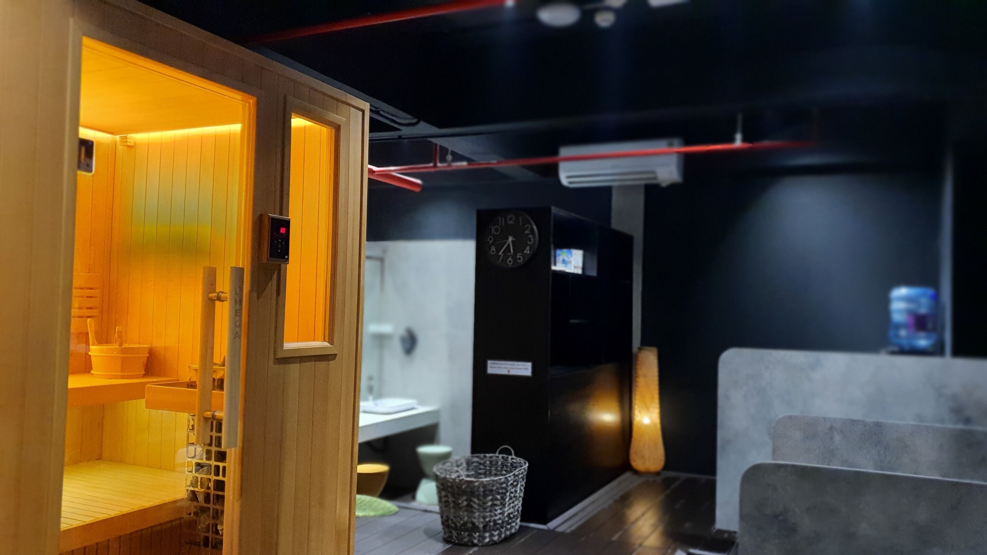 A sauna room with wooden walls and a bench, a digital clock, and a small basket on the floor.