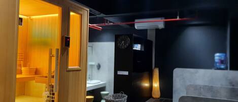 A sauna room with wooden walls and a bench, a digital clock, and a small basket on the floor.