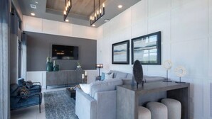 Una sala de estar moderna con un sofá gris, una mesa de centro y un televisor montado en la pared. Hay dos cuadros enmarcados en la pared, una mesita de noche con una lámpara y un objeto decorativo sobre la mesa de centro.