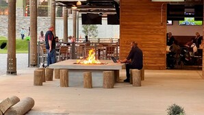 Un área de estar al aire libre con una chimenea central, pilares de madera y un muro de piedra al fondo.