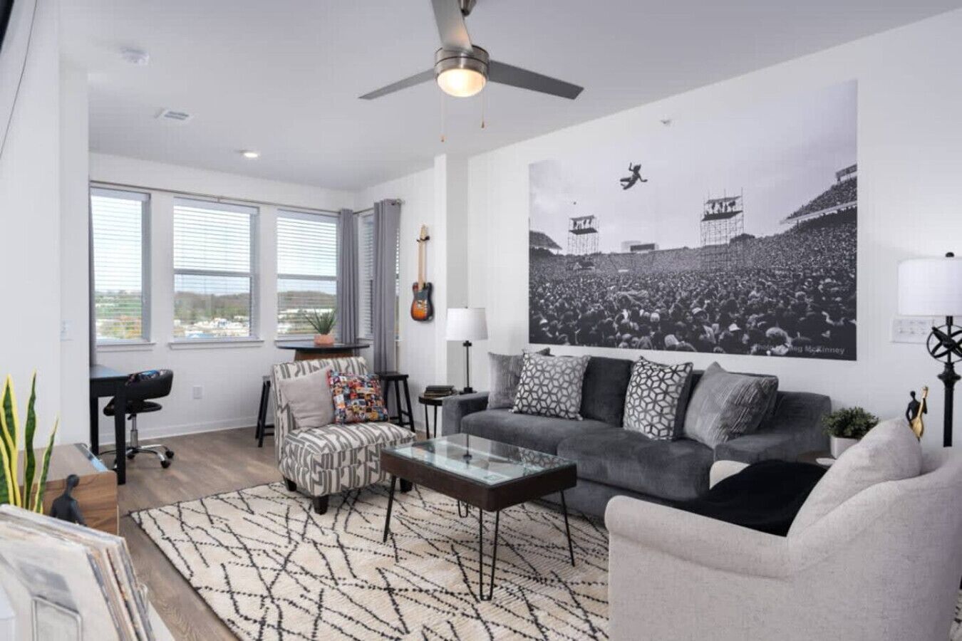A modern living room with a grey sofa, a patterned armchair, a glass coffee table, and a large black and white concert poster on the wall.