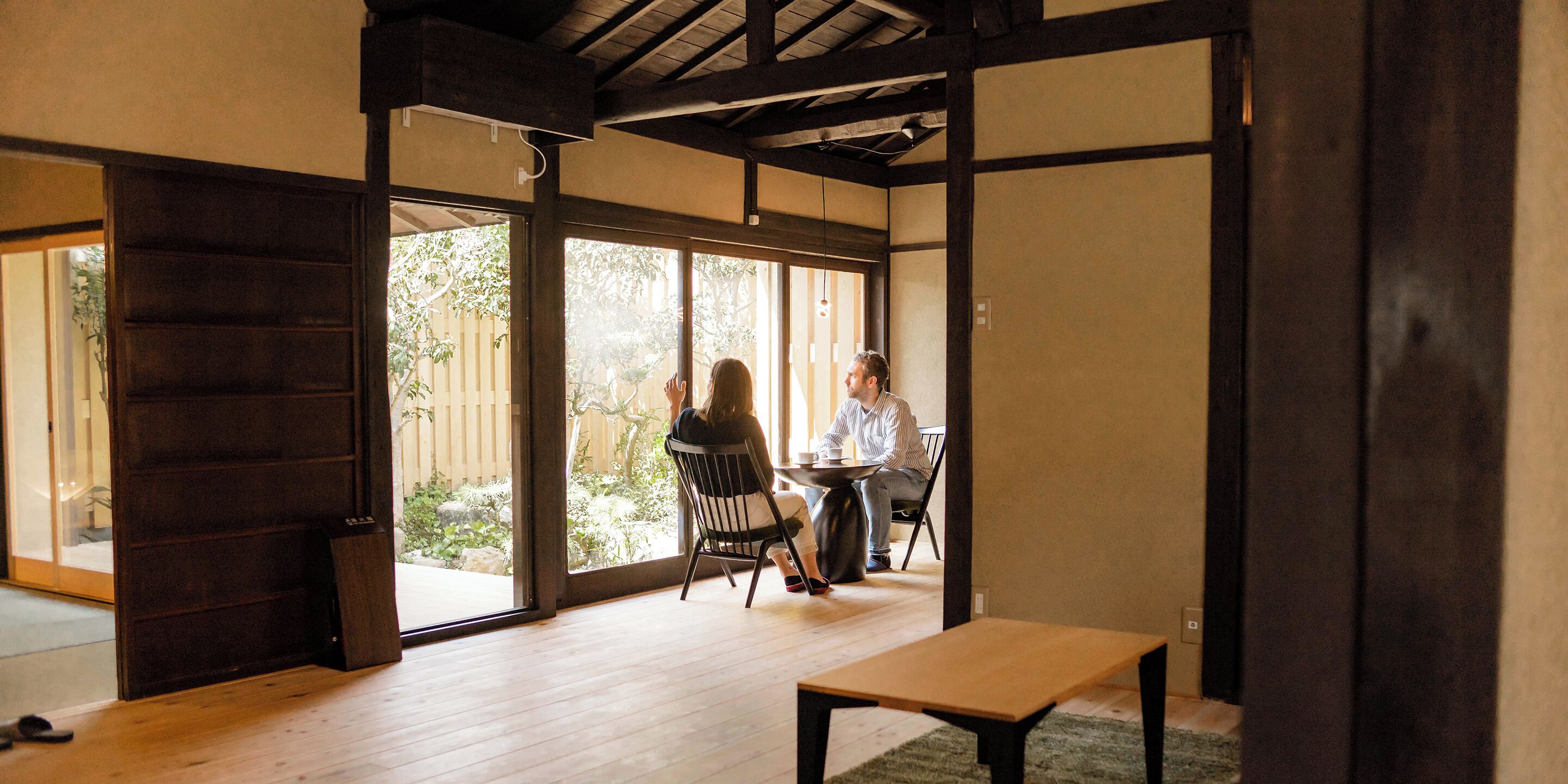A room with a wooden floor, a table, and a bench. There is a sliding door leading to an outdoor area with a view of trees and a fence.