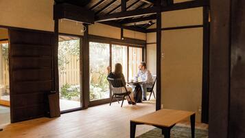 A room with a wooden floor, a table, and a bench. There is a sliding door leading to an outdoor area with a view of trees and a fence.