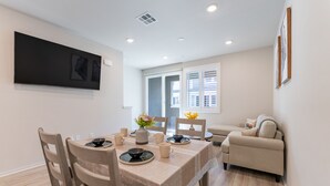 A dining area with a table set for four, surrounded by wooden chairs. There is a sofa in the background and a flat-screen TV mounted on the wall. - Brand New 3b3b Condo in the heart of West Covina (West Covina)