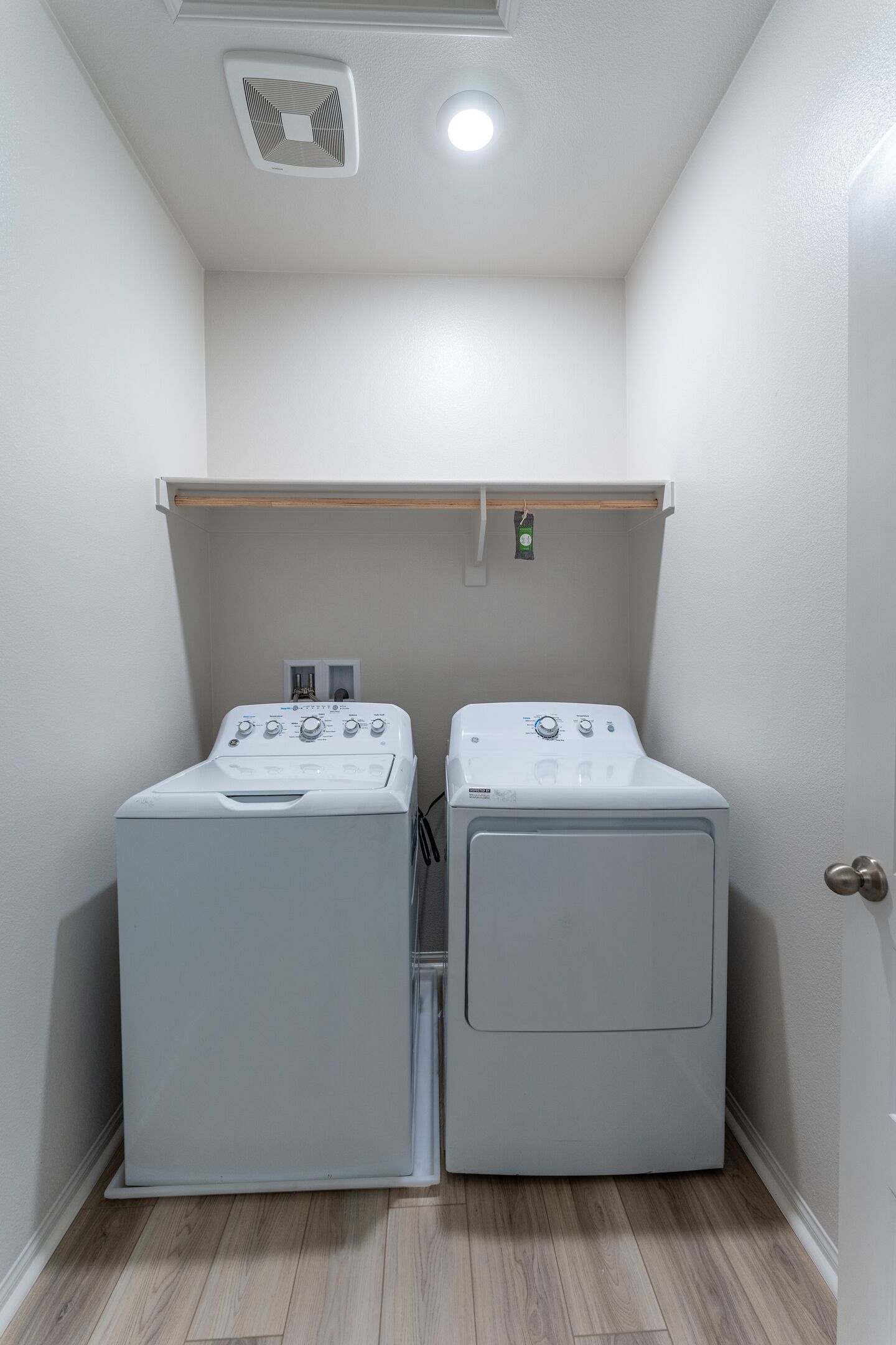 A laundry room with two white front-loading washing machines and a wooden shelf above them.