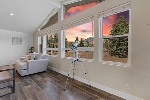 A room with a telescope on a tripod, a sofa, and large windows with a view of trees and mountains.