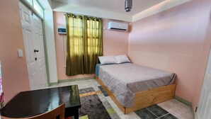 Un dormitorio con cama, escritorio, silla, ventana con cortinas, aire acondicionado y una lámpara en el techo.