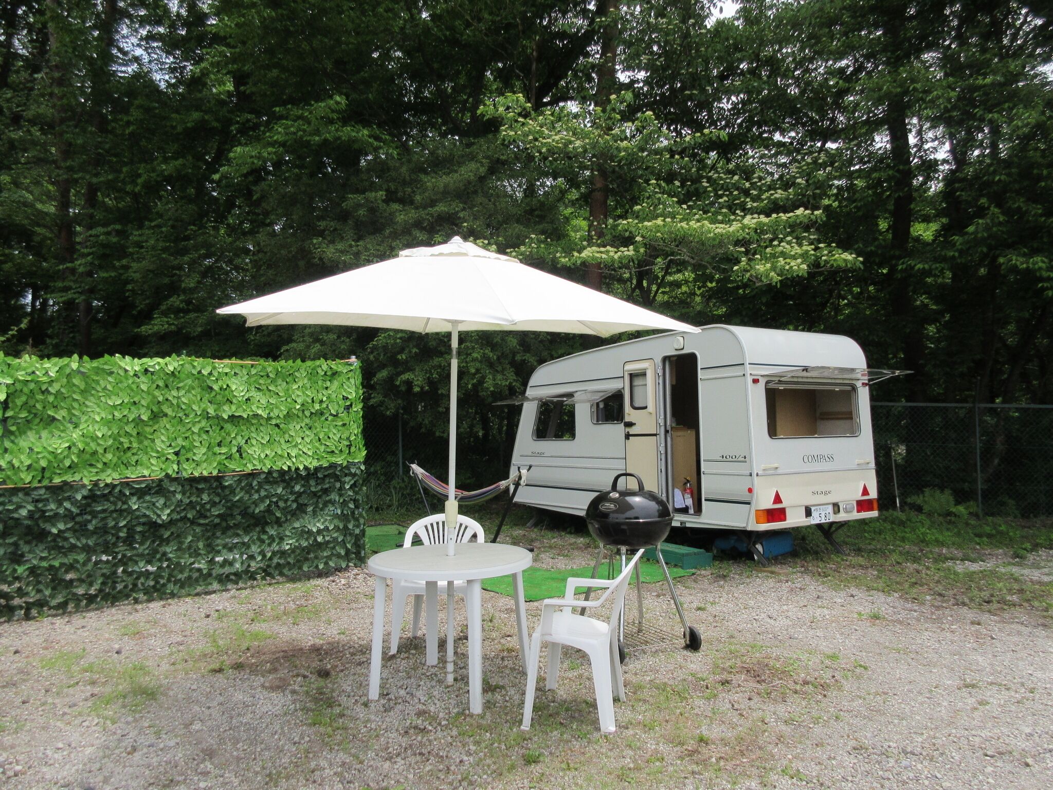 A camping setup with a white table and chairs under a canopy, a white camper with a grill, and a hedge in the background.