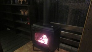 Ein Kaminofen mit Feuer, auf einem Fliesenboden stehend, mit einer hölzernen Bank und einem Regal mit Gegenständen im Hintergrund.