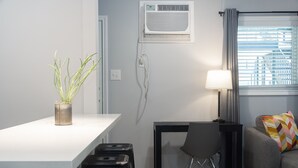 Ein modernes Wohnzimmer mit einem weißen Tisch, einem schwarzen Sessel, einem grauen Sofa mit farbigem Kissen, einer Topfpflanze und einem Fenster mit Rollläden.