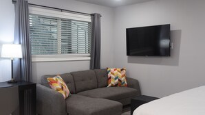 Ein modernes Hotelzimmer mit grauem Ecksofa, einem flachbildfernseher an der Wand, einem Nachttisch und einem Fenster mit Jalousien.