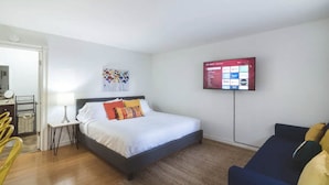 Habitación de hotel con cama, televisión montada en la pared, una mesita de noche con lámpara y una silla.