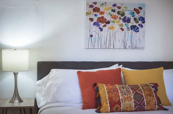 A bed with white and colorful pillows, a lamp, and a wall with a floral painting.