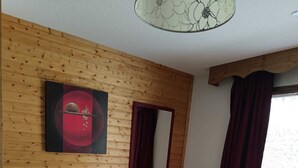 Ein Wohnzimmer mit grauem Sofa, Holzwandverkleidung, einem großen Fenster mit roten Vorhängen und einer Deckenleuchte.