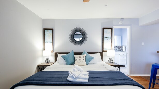 A neatly made bed with a dark blue bedspread and white linens, two nightstands with lamps, a wall mirror, and a view into a kitchen area.