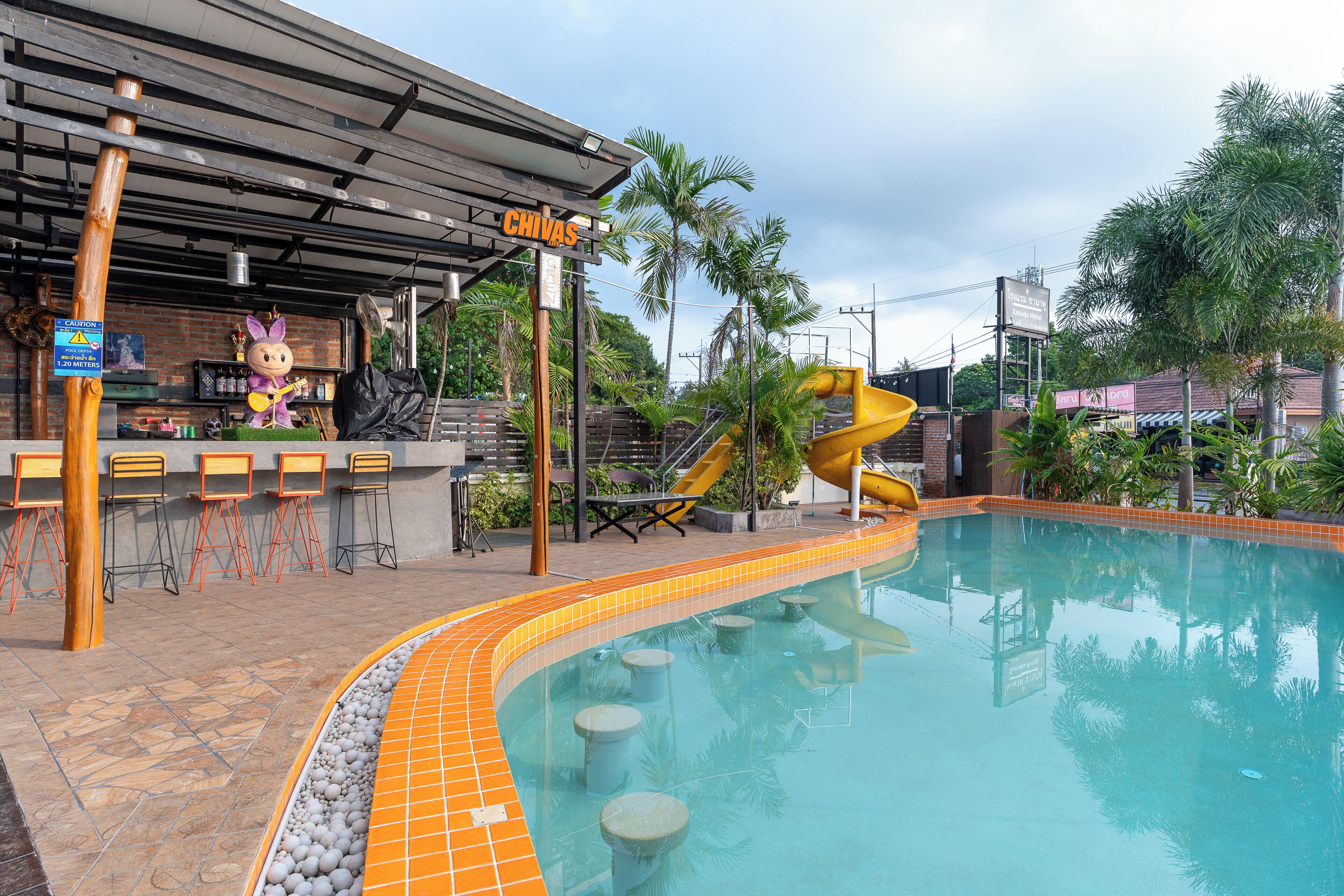 Zona de piscina con tobogán, bar con un personaje representativo y asientos al aire libre.