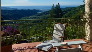 A balcony with a view of a mountainous landscape, a chair, and a table with a book.