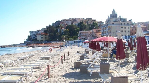 A beach with sun loungers and umbrellas, a lifeguard tower, and buildings in the background.