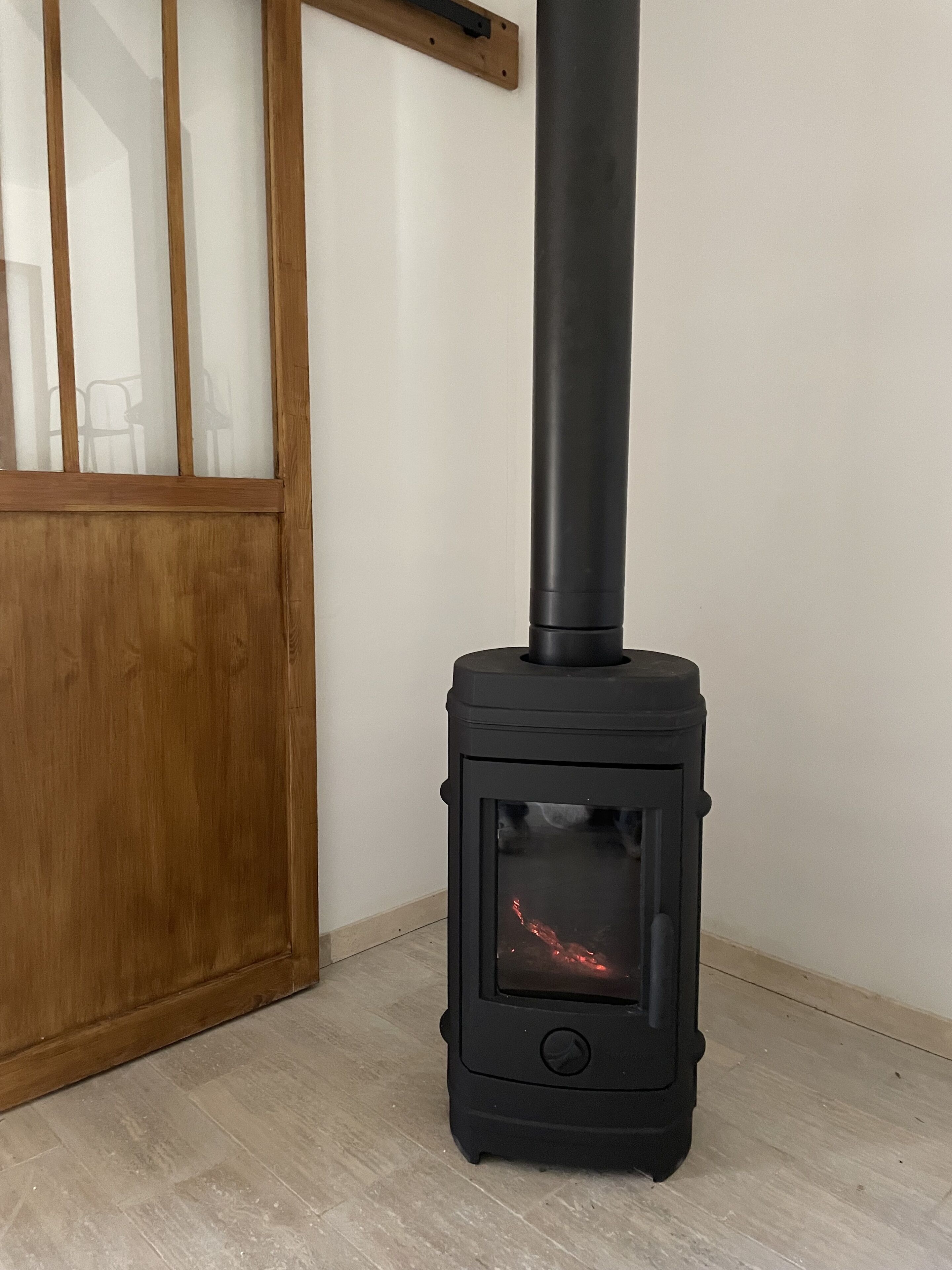 A black wood-burning stove with a glass door, positioned in a corner of a room with wooden flooring and a wooden door.