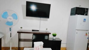 Une petite pièce meublée, avec un téléviseur à écran plat fixé au mur, un réfrigérateur blanc, une table blanche sur laquelle se trouvent une bouteille et des mouchoirs, un ventilateur sur pied et une petite plante posée sur une étagère.