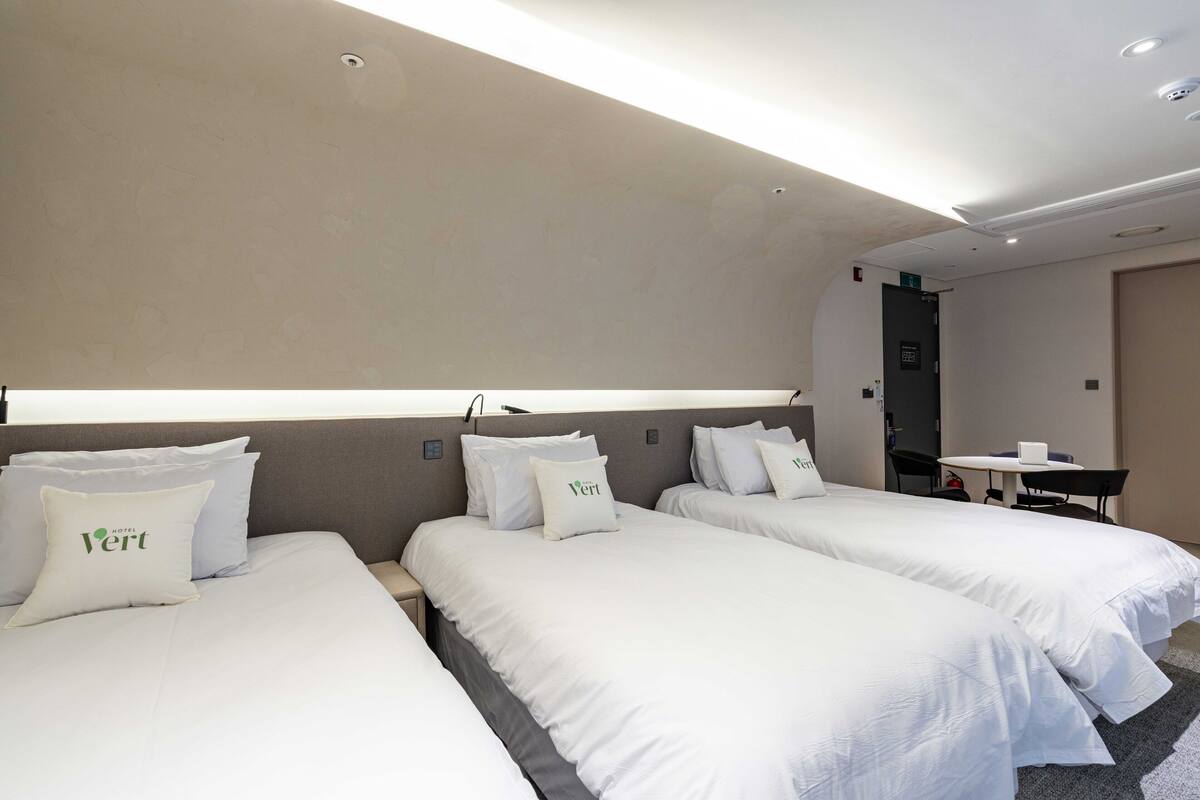 A hotel room with three beds, each with white bedding and pillows, a small table, and a chair.