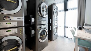 A laundry room with stacked washing machines and dryers, a table with chairs, and a large window with curtains. - Hotel Vert (Seoul)