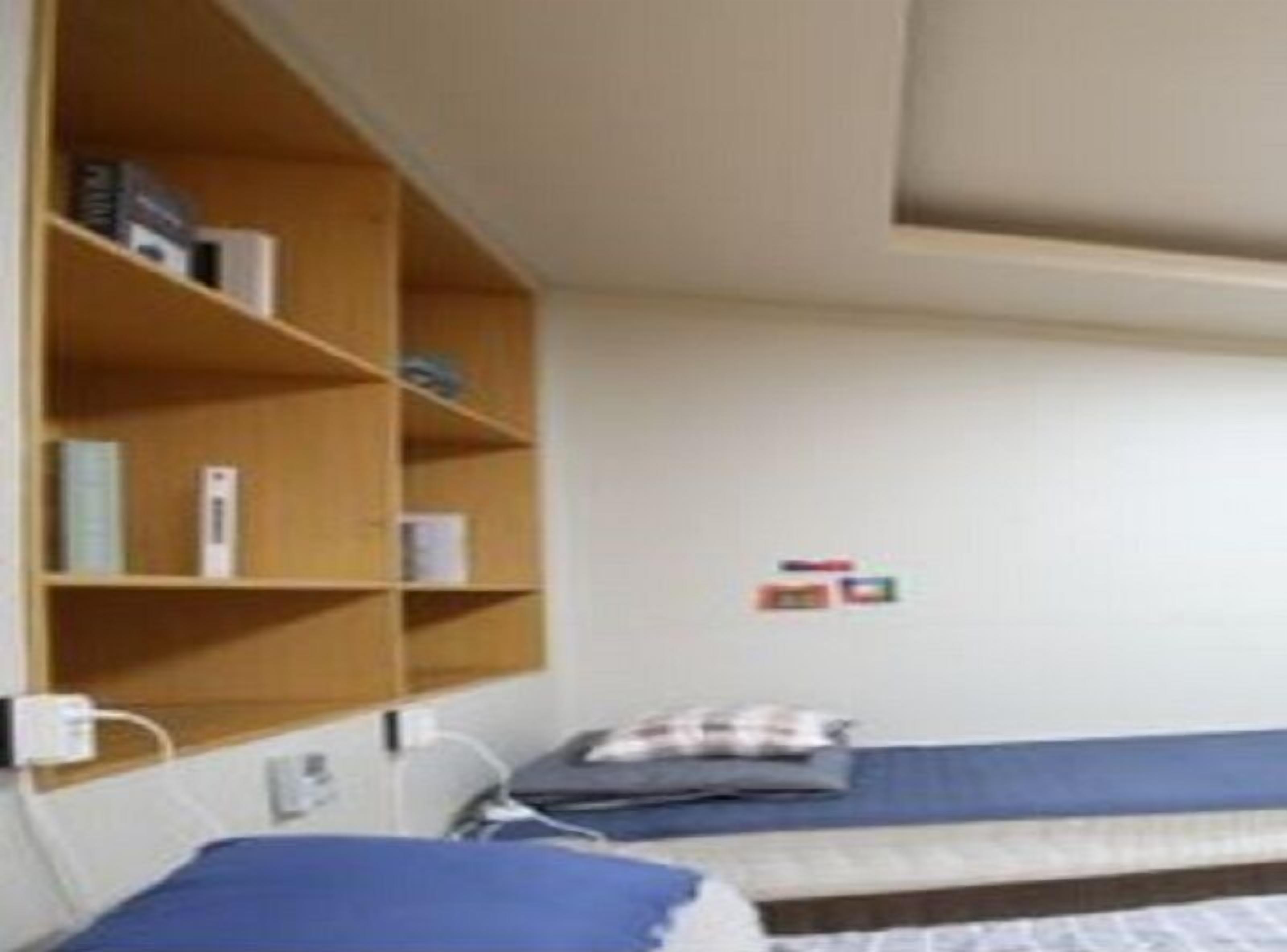 A single-bed room with a wooden bookshelf, a bedside table, and a wall-mounted power outlet.