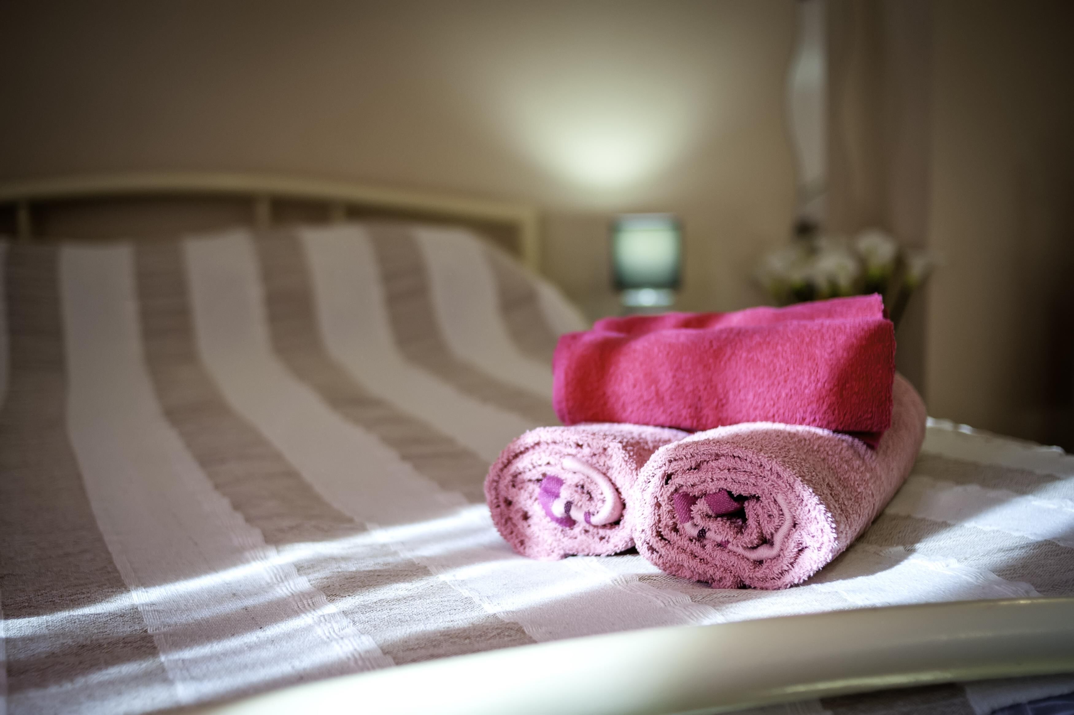 A bed with a striped bedspread, a rolled pink towel, and a small vase with flowers on the side.
