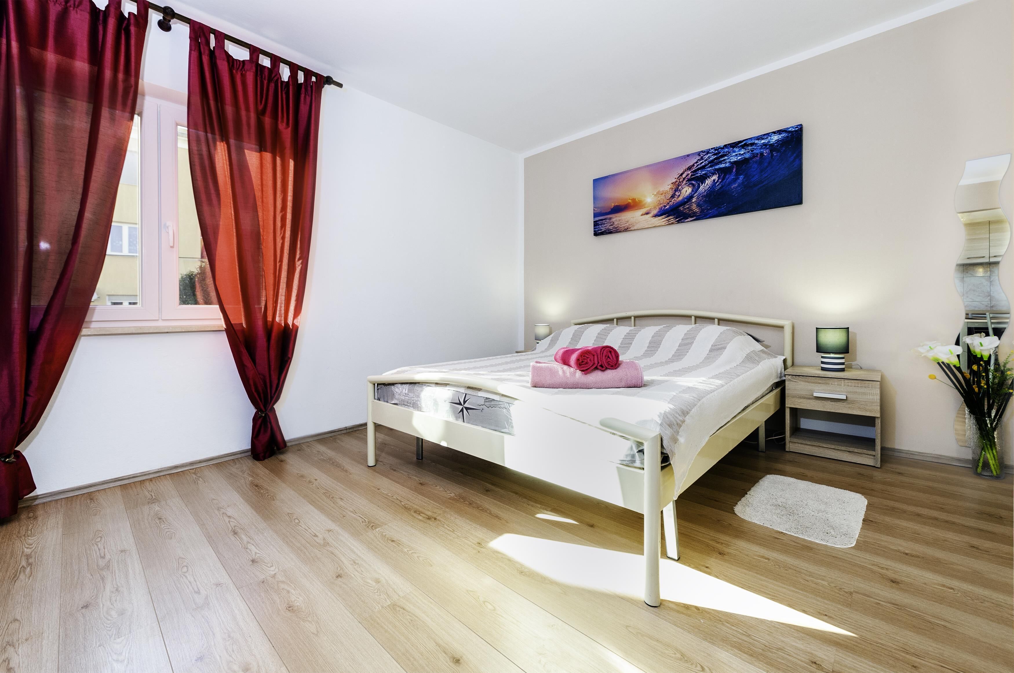 Une chambre à coucher avec un lit, des rideaux, une table de chevet, une commode, un tableau accroché au mur et une fenêtre.