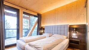 A bedroom with a large bed, a nightstand with a lamp, and a view of the outdoors through a sliding door. - Natural chalet with sauna and hot tub (Turrach)