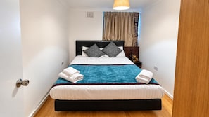 A neatly made bed with white linens, a blue blanket, and two pillows. There is a small bedside table with a lamp and a golden statue. The room has white walls, a wooden floor, and a window with curtains.