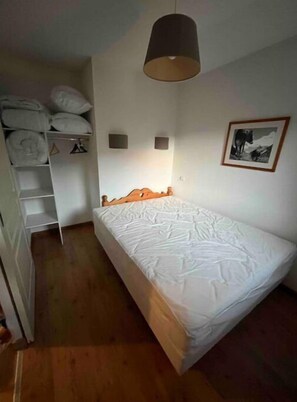 Una habitación pequeña con una cama individual, cabecera de madera, lámpara colgante, colcha blanca, armario blanco y un cuadro en la pared.