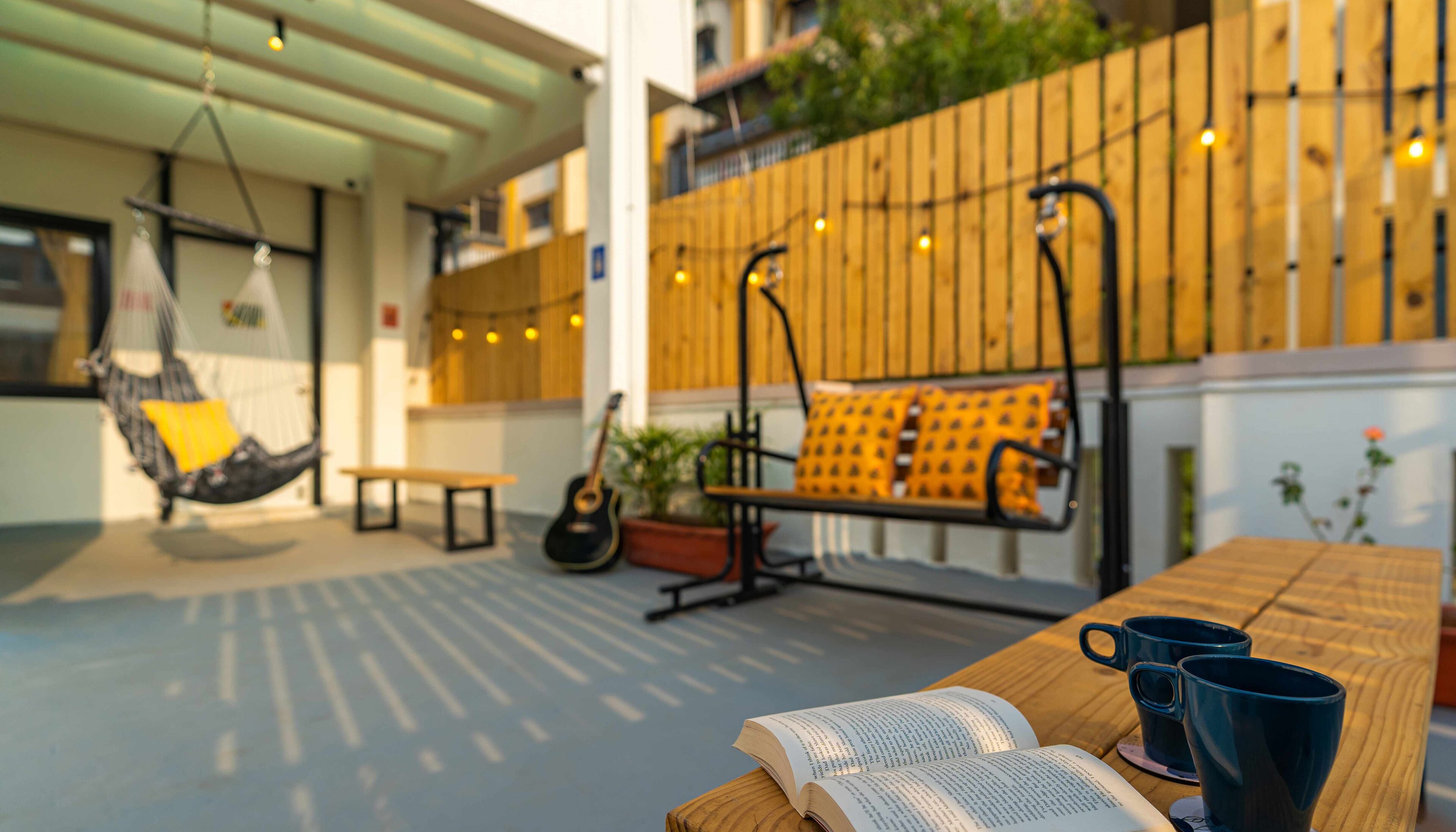 Un espace extérieur accueillant avec un hamac, un banc et une table en bois sur laquelle sont posés un livre et deux tasses.