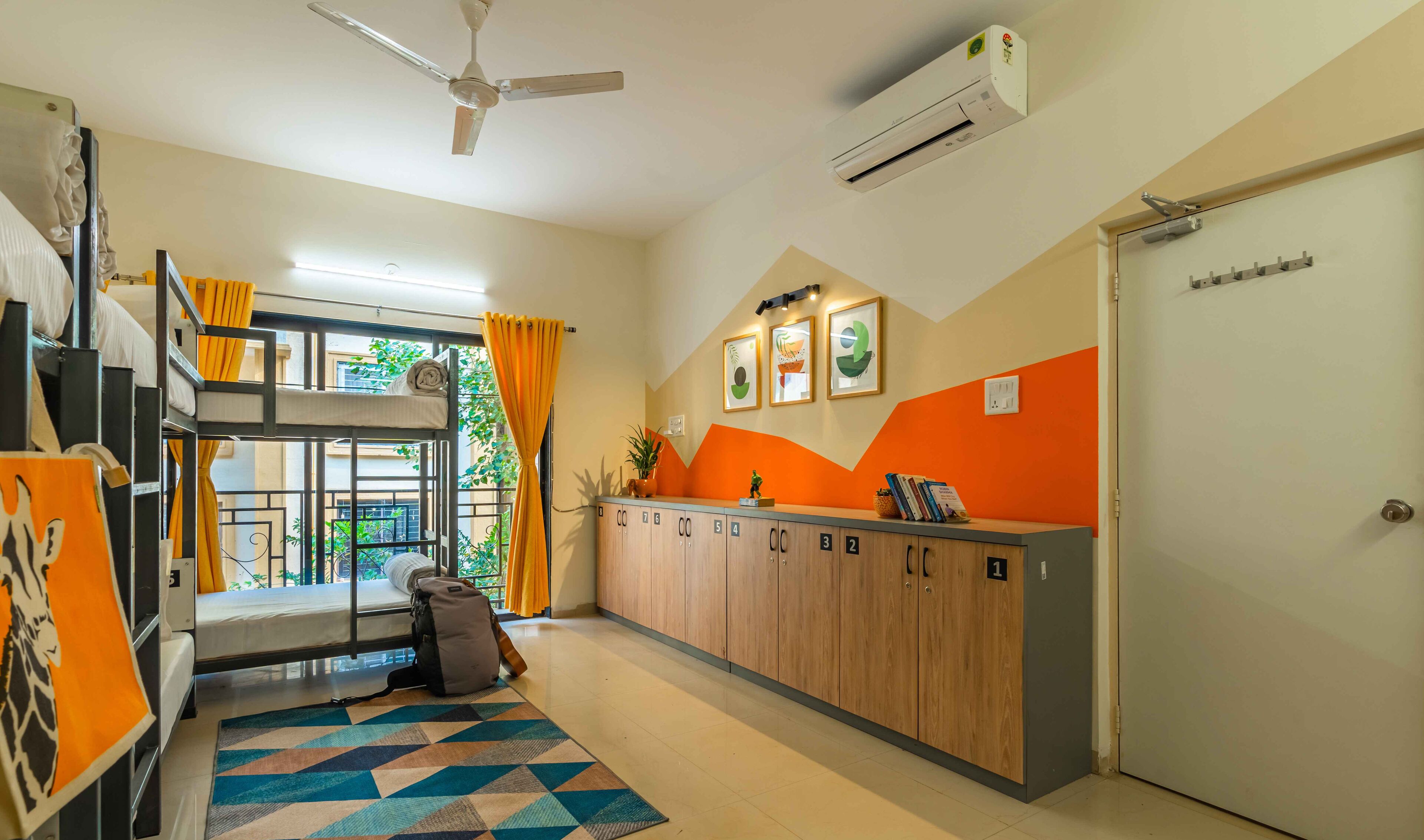 A dormitory room with bunk beds, a desk with lockers, a ceiling fan, and an air conditioning unit.
