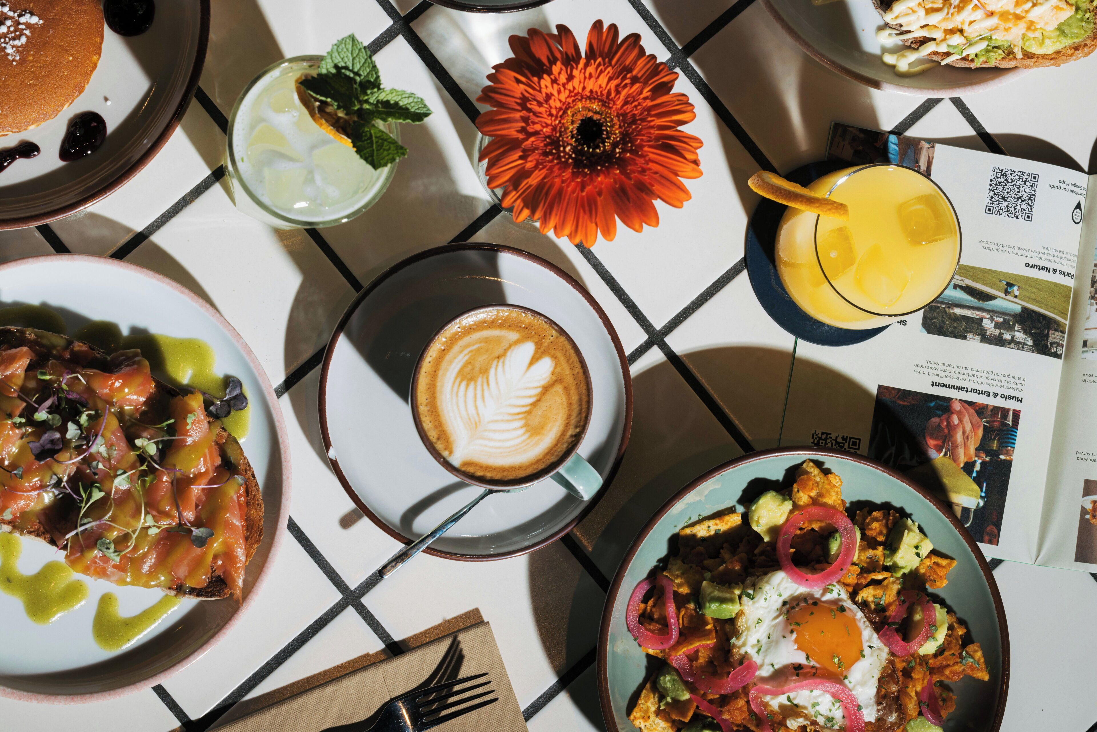 Et bord med morgenmad, en kaffekop med latte-kunst, en blomst og en menu.