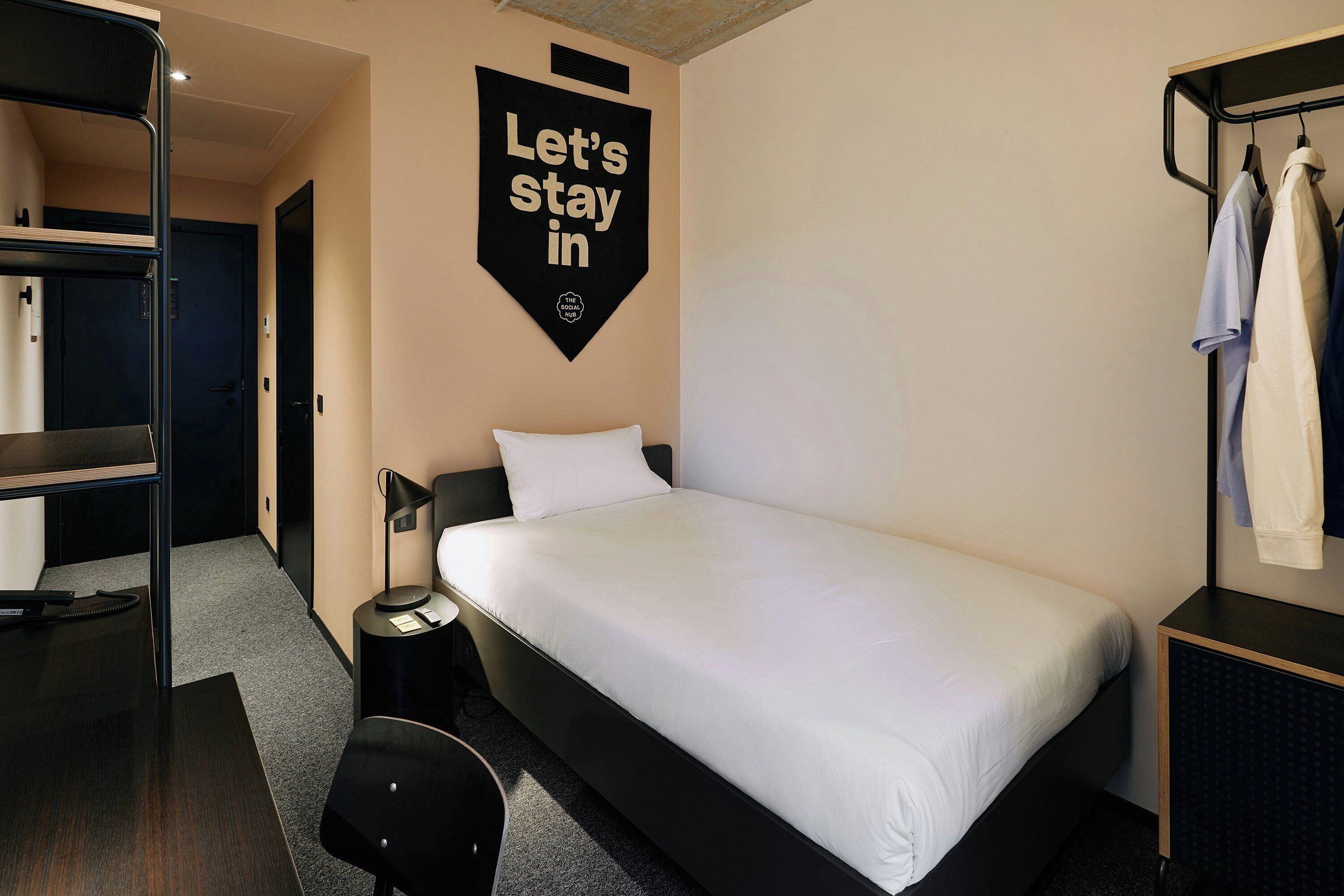 A hotel room with a bunk bed, a single bed, a desk with a chair, a bedside table, and a wall-mounted towel rack.