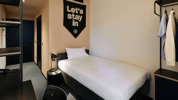A hotel room with a bunk bed, a single bed, a desk with a chair, a bedside table, and a wall-mounted towel rack.