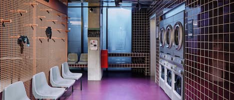 A public laundromat with rows of washing machines, a wall-mounted drying rack, and a seating area with chairs.