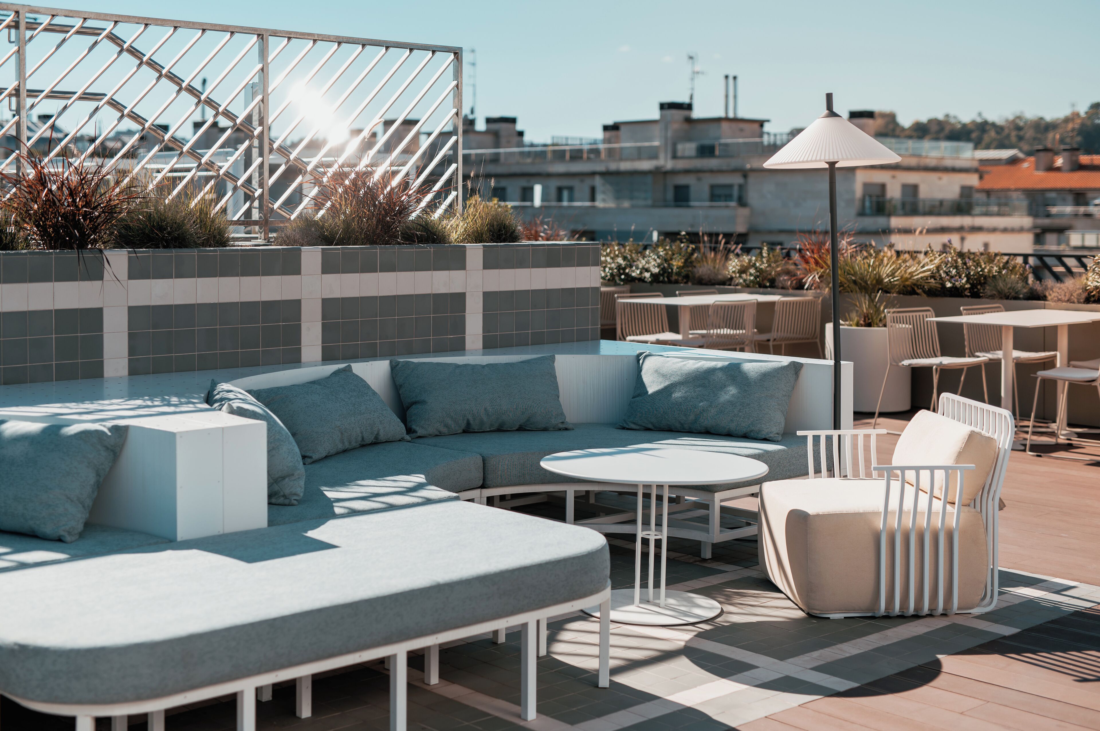 A rooftop terrace with a sectional sofa, round tables, and chairs, surrounded by potted plants and a metal railing.