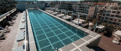 A rooftop pool with clear blue water, surrounded by lounge chairs and sunbeds, overlooking a cityscape with buildings and mountains in the distance.