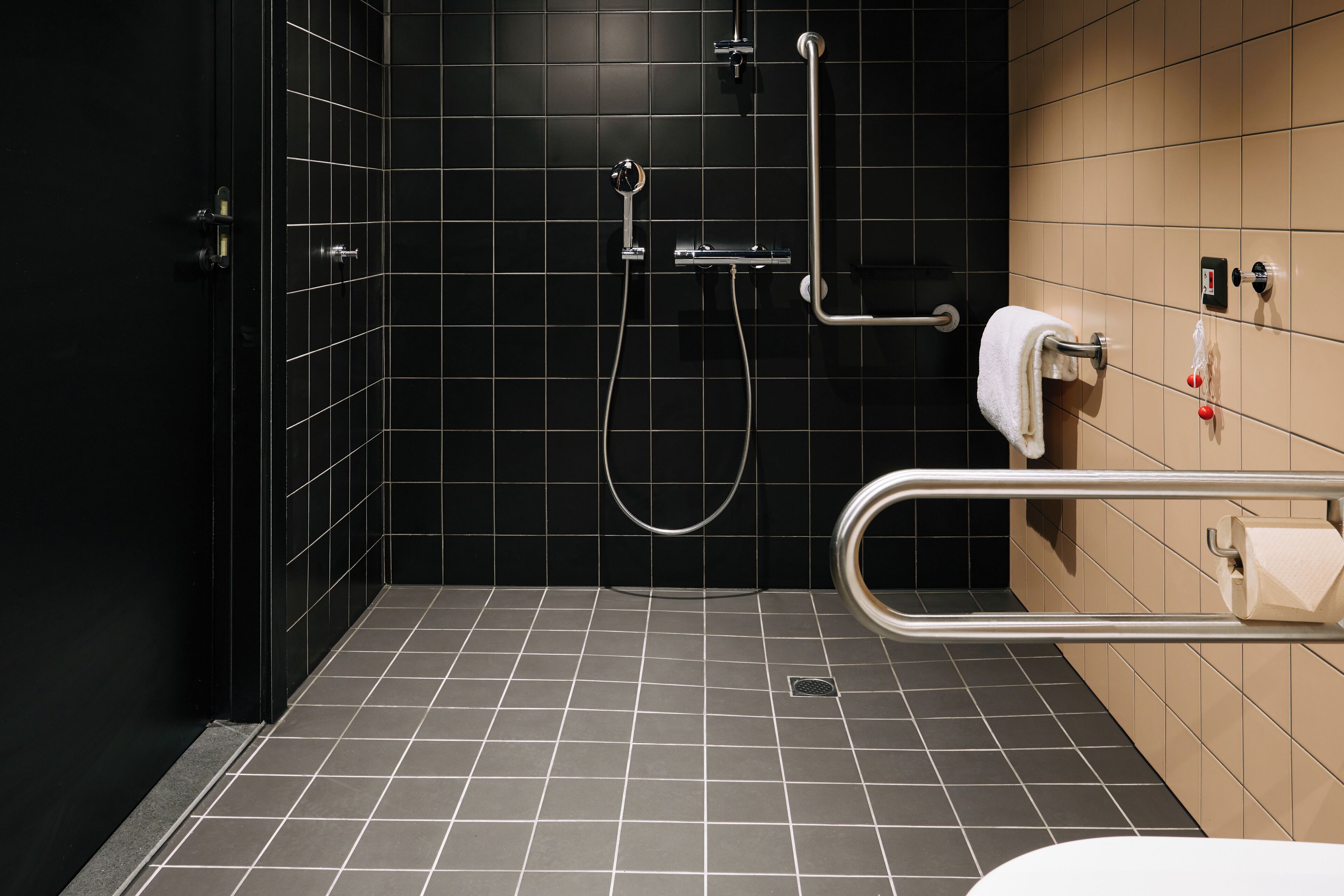 Ein modernes Badezimmer mit schwarzen Fliesenwänden, einer Dusche inklusive Handbrause, einer Haltestange und einem Handtuchhalter.